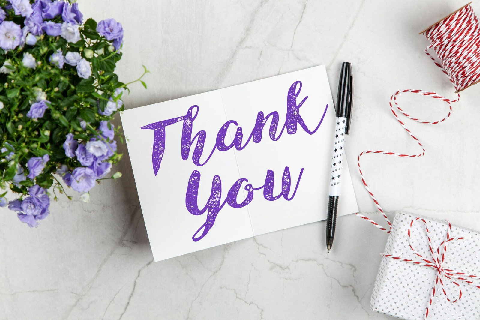 A stylish thank you card paired with purple flowers, a gift box, and pen on a marble surface.